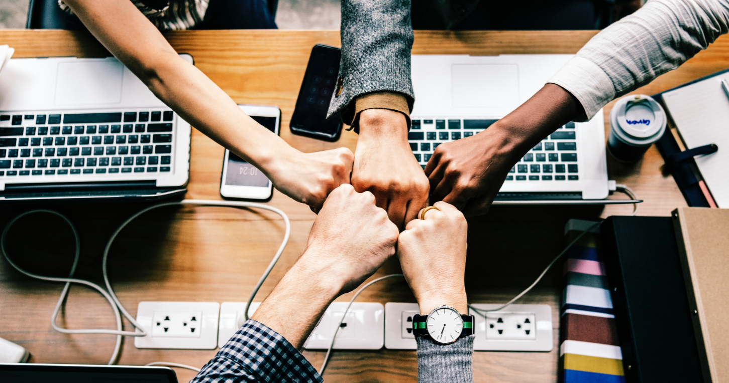 5 Hände von 5 Personen über einem Konferenztisch zusammengehalten